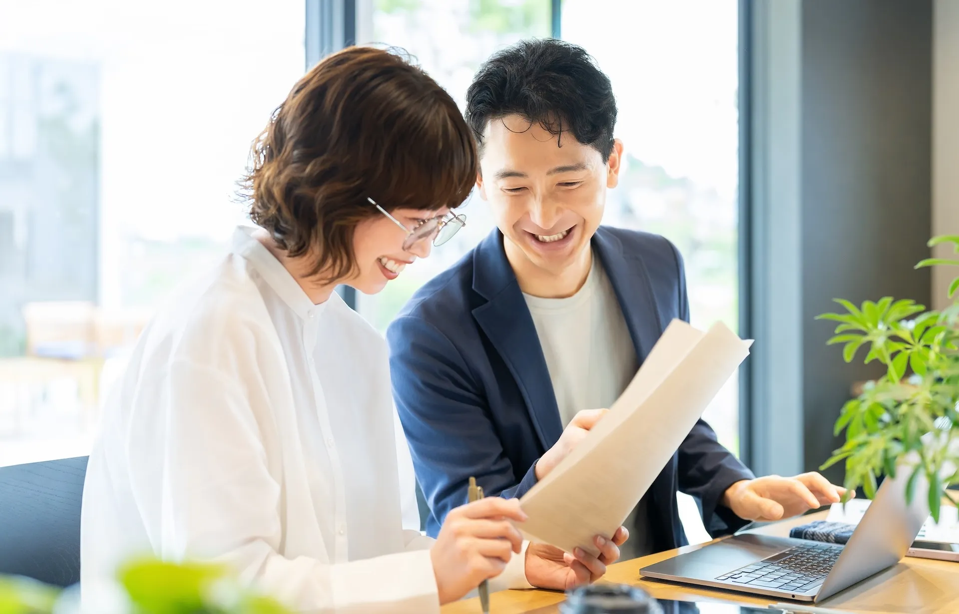 Two coworkers smile while reviewing documents at a bright office with a laptop on the desk.